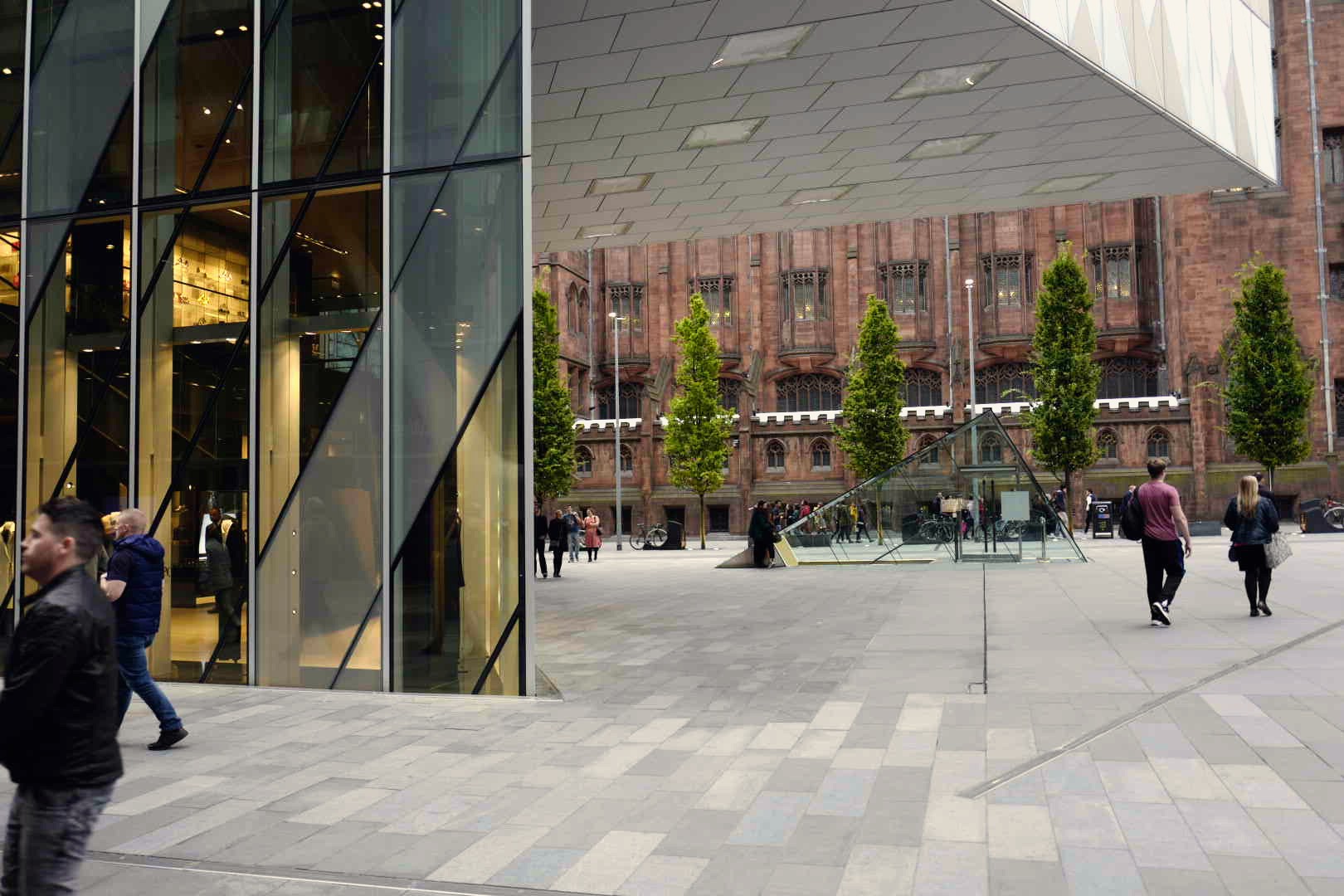 Pedestrians crossing a paved public square beside a modern glass building in central Manchester.